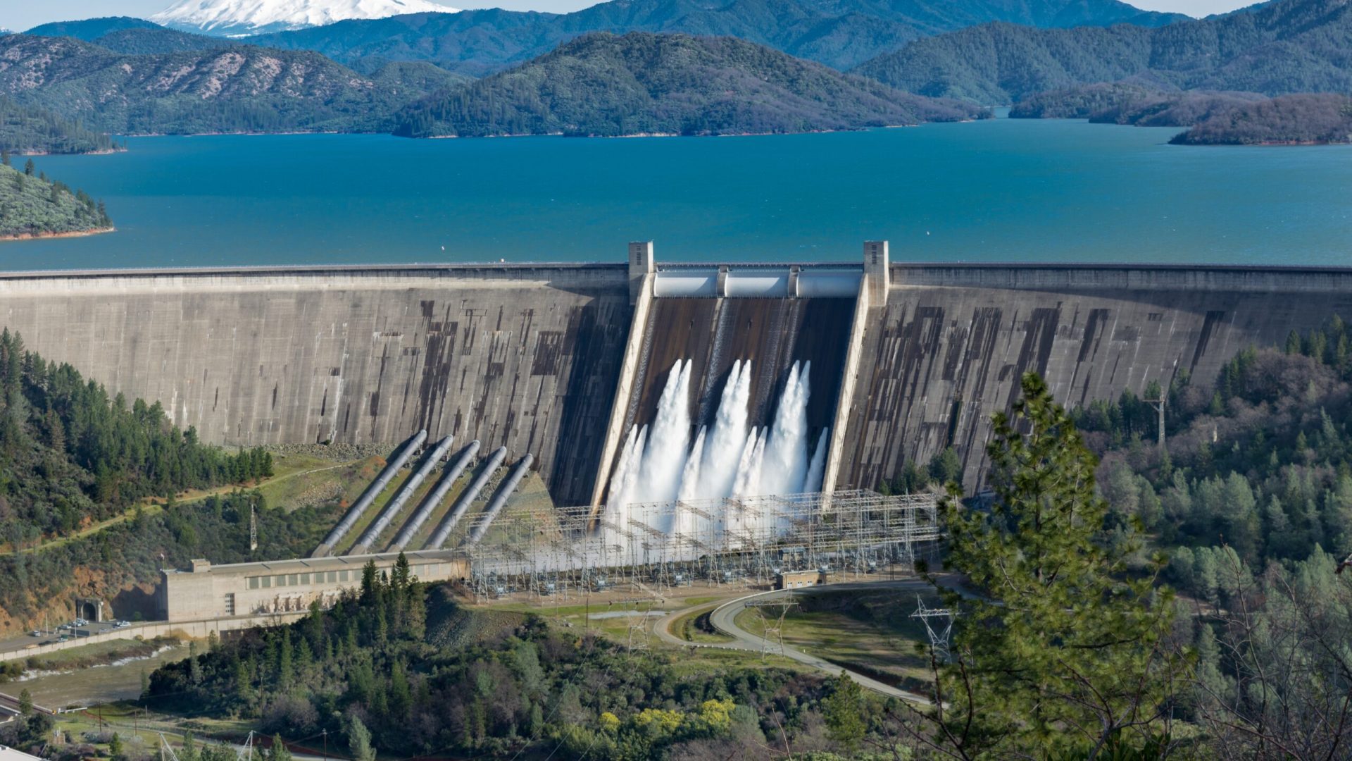 A picture of Shasta Dam surrounded by roads and trees with a lake and mountains on the background